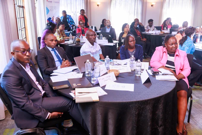 A group of people seated at round tables in a conference room attentively listen to a presentation. The setting is professional, with notepads, laptops, and water bottles on the tables. In the background, other attendees are visible near banners displaying the "FHI 360" and "USAID" logos.