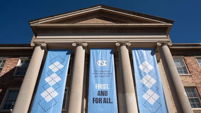 Facade of a historic building with columns, displaying two large blue banners. The banners read "The University of North Carolina at Chapel Hill" with "First. And For All." underneath. The university's logo is also visible.