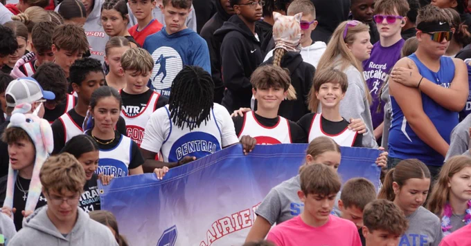 A diverse group of young athletes in sports uniforms holding a large blue banner during an outdoor event.