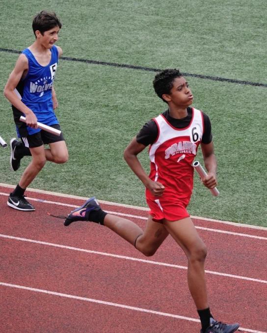 Two young athletes sprinting on a track during a relay race, each holding a baton and wearing numbered jerseys.