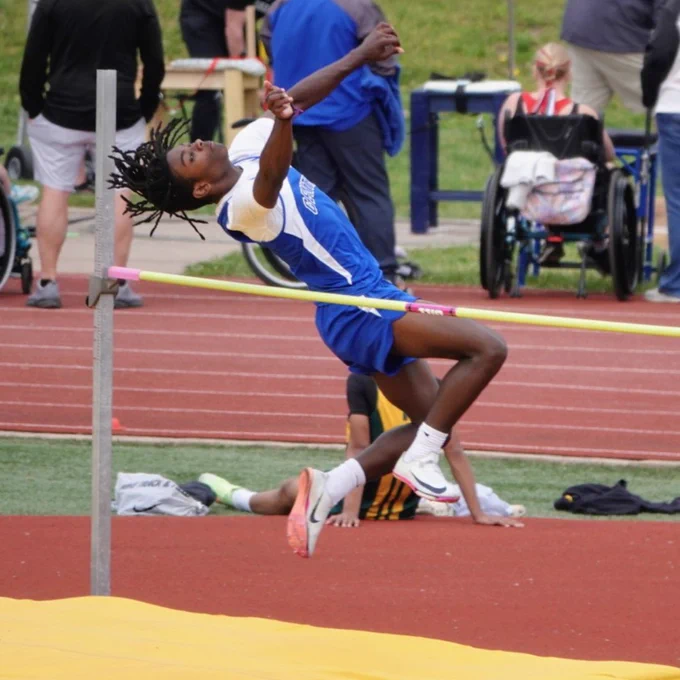 Athlete in blue uniform performing a high jump over the bar on a track and field stadium.