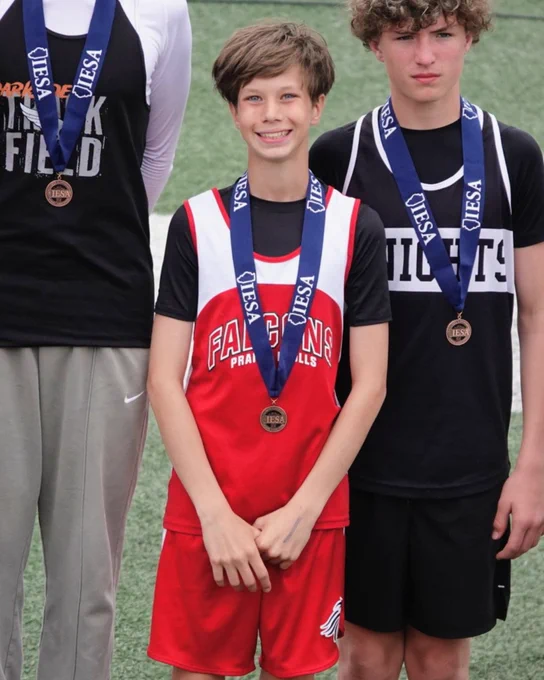 Three young athletes wearing medals on blue ribbons stand side by side on a sports field, dressed in different team uniforms.