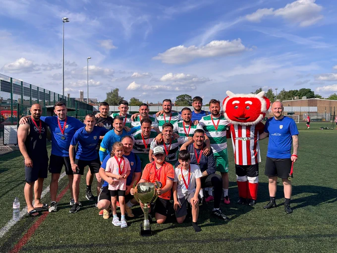 Group of footballplayers and children posing on a field with a large trophy and a red mascot in a striped jersey under a sunny sky.