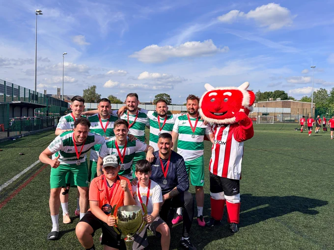 Football team in green and white hooped uniforms posing with a large trophy and a red mascot on a sunny outdoor field.