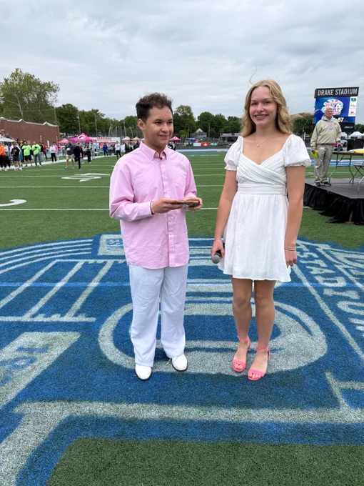 Two young people standing on a football field logo, dressed in semi-formal attire with the boy in a pink shirt and white pants, and the girl in a white dress and pink heels.