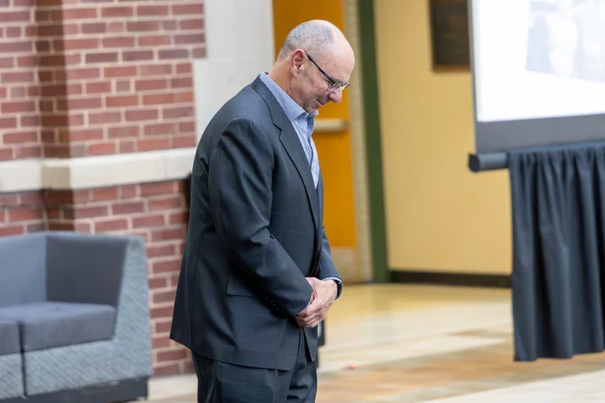 A person in a suit bows slightly in a professional indoor setting. The mood is respectful and contemplative.