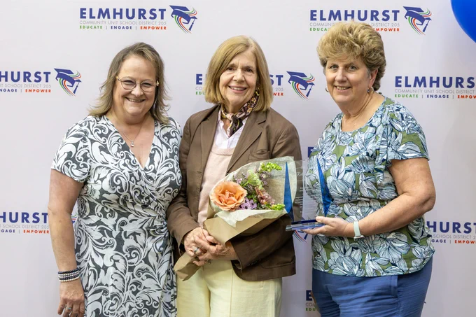 Three women stand together at an Elmhurst Community School District 205 event, one holding flowers and another holding an award. The mood is celebratory.