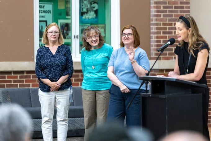 A group of women stands next to a podium with a speaker addressing an audience. The setting appears to be a formal indoor event.