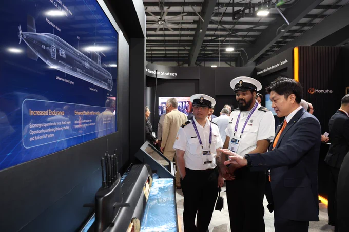 Three men, including two in naval uniforms, discuss a submarine display at a technology exhibition booth.