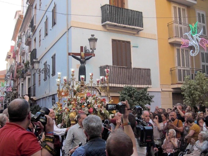 Procesión religiosa en calle: paso floral con crucifijo, rodeado de fotógrafos y público observando.
