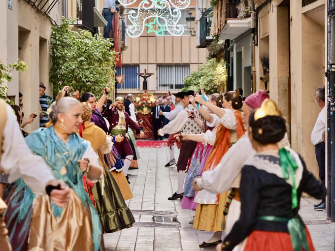 Danza tradicional en calle estrecha: filas de bailarines y procesión con crucifijo y carro floral.