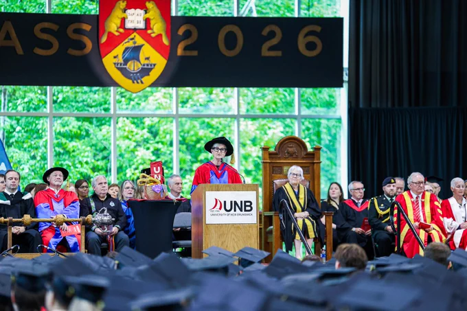 A graduation ceremony at the University of New Brunswick features several dignitaries in academic regalia on stage. A person stands at a podium displaying the UNB logo and addresses the audience. Behind the speaker, a sign reads "Class 2026," and the university crest is displayed. Graduates in caps are seated in the foreground.