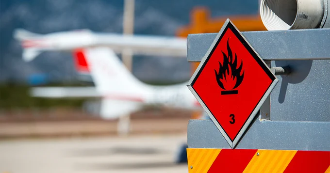 A red flammable materials warning sign with a flame symbol is displayed prominently. In the background, a plane is visible on an airfield.
