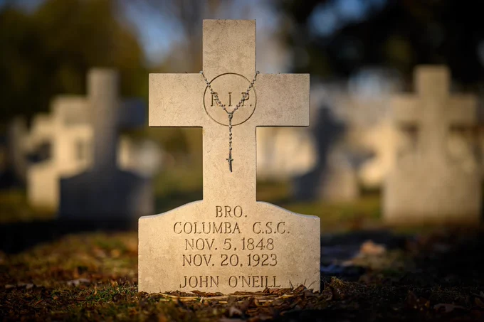 A cemetery scene with a stone cross headstone in the foreground. The headstone has "R.I.P." and a rosary engraved at the top, followed by "Bro. Columba C.S.C. Nov. 5, 1848, Nov. 20, 1923," and the name "John O'Neill" at the bottom. The background shows more blurred headstones.