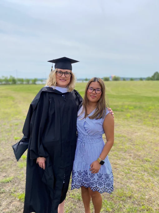 A woman in a black cap and gown stands smiling beside a young woman in a blue-and-white striped dress in an open grassy field.