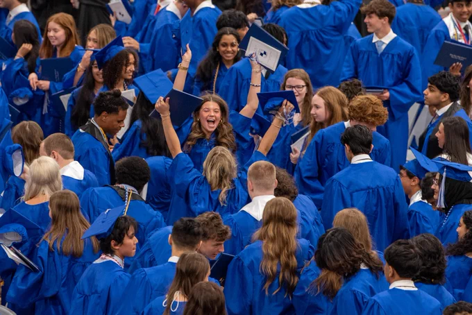 Large group of people wearing blue robes and hats, many with arms raised excitedly, gathered in an auditorium or stadium.