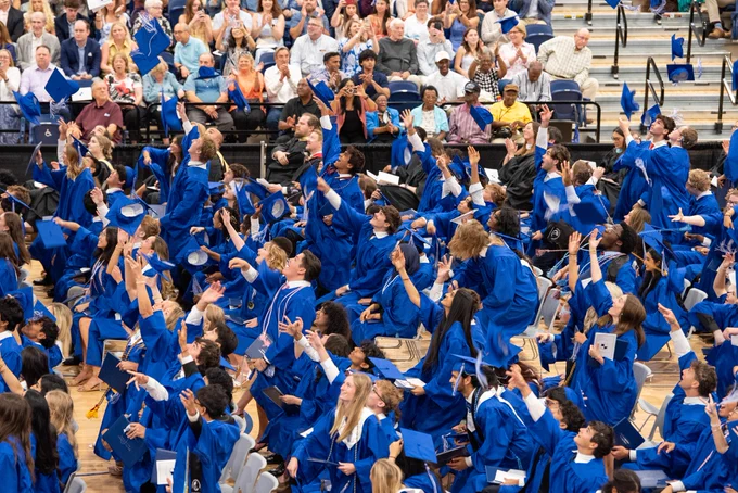 A crowded stadium filled with people in blue graduation gowns and caps, with some waving and cheering.