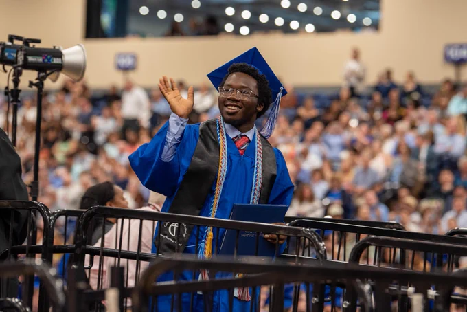 Smiling graduate in blue robe and cap, waving from stage at graduation ceremony.