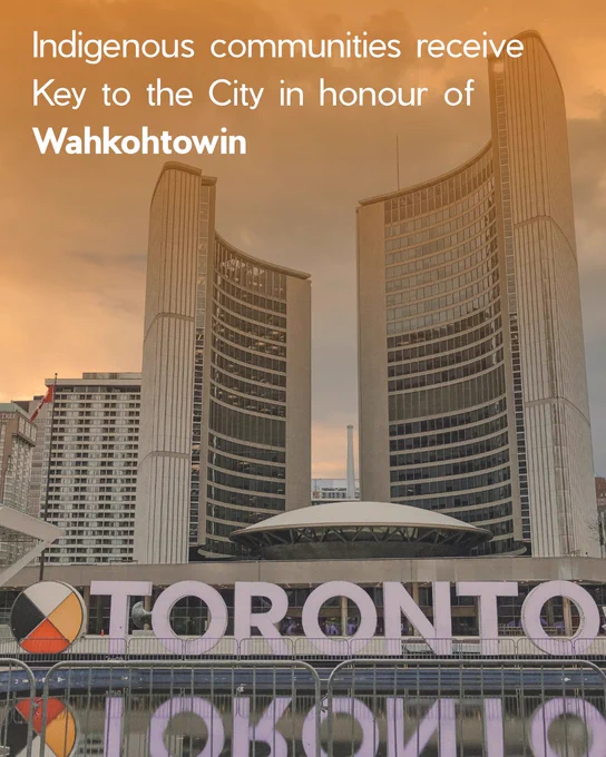 A wide view of Toronto City Hall showing the two curved concrete towers rising behind the circular council chamber. In the foreground, large letters form the “TORONTO” sign in Nathan Phillips Square. A warm orange filter covers the photo. Overlaid text at the top of the image reads: “Indigenous communities receive Key to the City in honour of Wahkohtowin.”