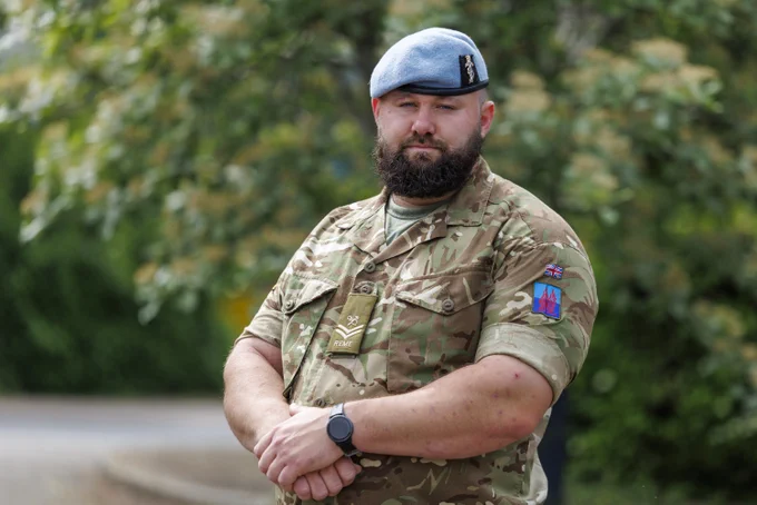 A person in a British Army uniform stands outdoors. The uniform features camouflage patterns and various insignias, including a Union Jack patch on the arm. The person is wearing a blue beret. Trees and greenery are visible in the background.