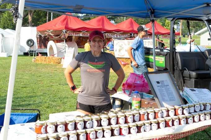 A person posing for a photo at the Iowa State Fair Farmers Market with a table full of Jam and Jelly. 