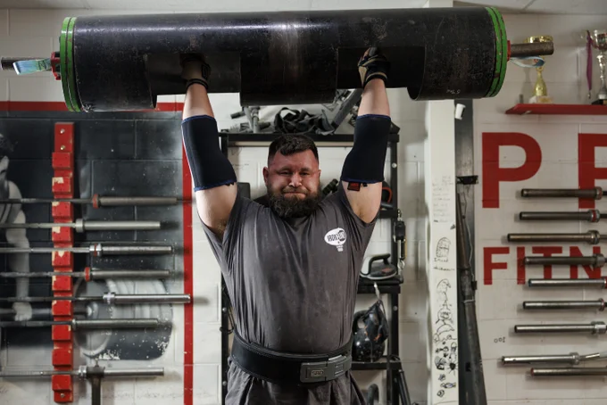 An individual is lifting a large, heavy log-shaped weight overhead in a gym. The gym has various weights and equipment visible, including a wall with "PF FITNESS" partially showing. The individual is wearing a dark shirt and wrist supports.