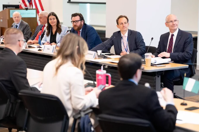 A group of individuals are seated at a conference table in a formal meeting room, with a focus on a row of six people facing the camera. Some participants are reviewing documents. The background features flags, including the American flag.