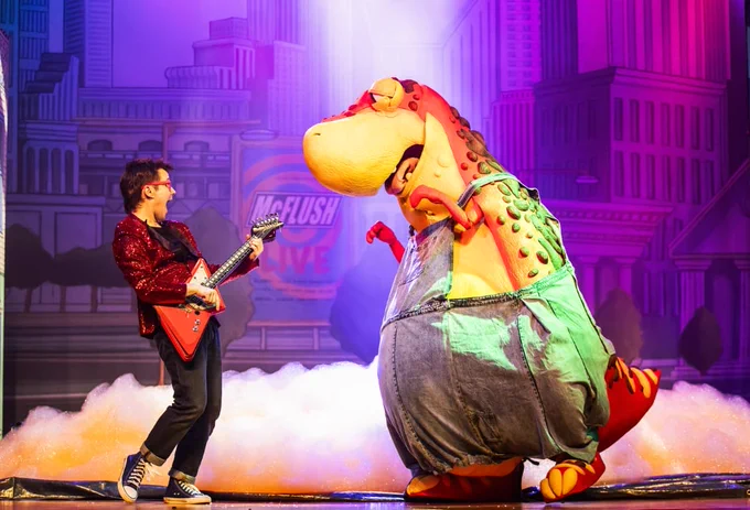 A person in a sparkling jacket plays an electric guitar on stage. Opposite, a large costumed character resembling Earl Sinclair from "Dinosaurs" stands in oversized clothing. The backdrop features a stylized cityscape with bright, colorful lights.