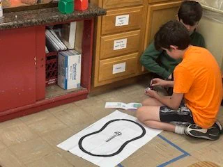 Two students sit on the classroom floor with their robot, attempting to get it to follow a line drawn on paper.  