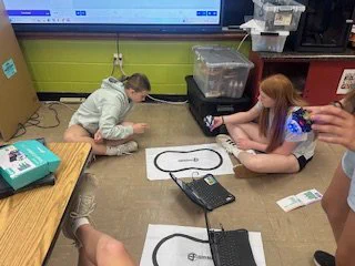 Two students sit on the classroom floor with their robot and laptop, attempting to get it to follow the line. 