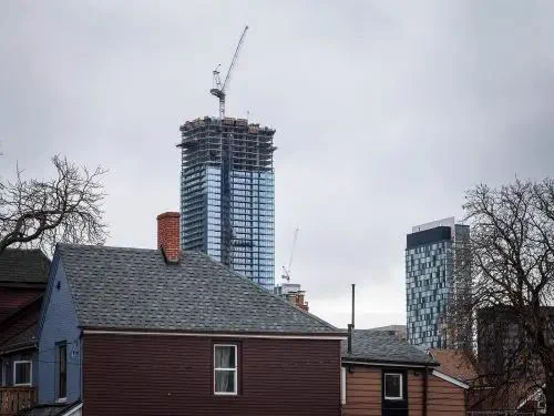A towering modern skyscraper under construction, topped with a crane, contrasts against overcast skies and low-rise residential homes in the foreground.