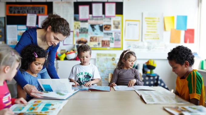 A group of young children sits around a table in a classroom, each engaged with books. A person is assisting one of the children. The walls are decorated with colorful educational materials and posters.