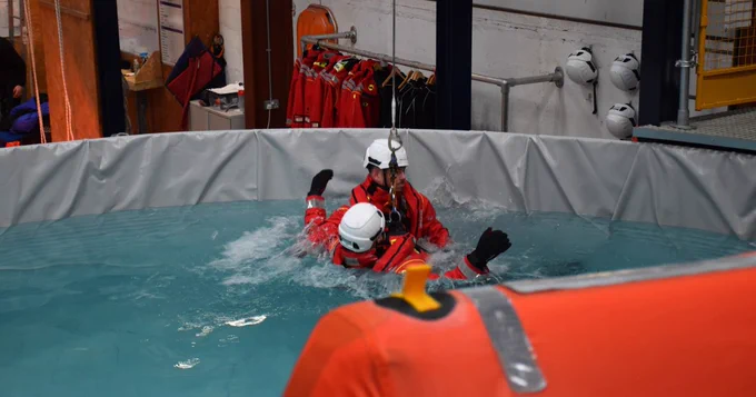 A participant being dropped into the sea survival tank from a winch and a person helping.