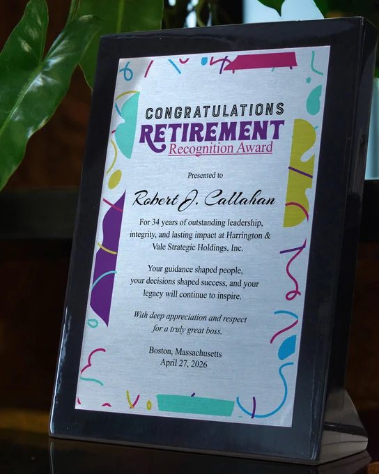 Custom Aluminum Award With Black Piano Finish And Printed Retirement Message Standing  Via Easel On Wood Shelf With Plant