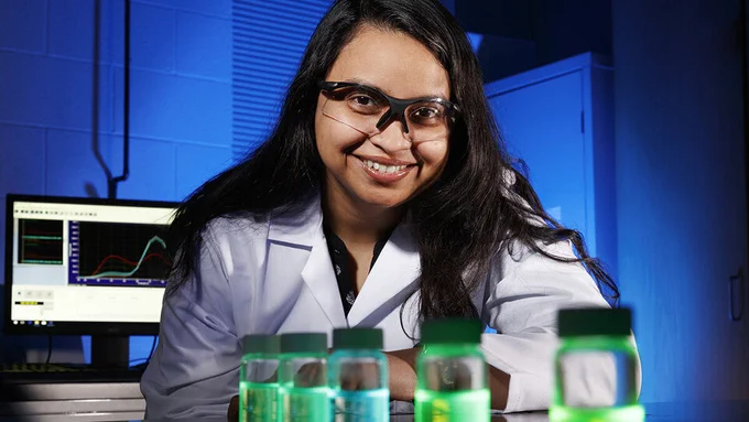 Shudipto Dishari leaning against a lab counter. She is in a lab coat and goggles, with vials of liquid in front of her and a computer behind her.