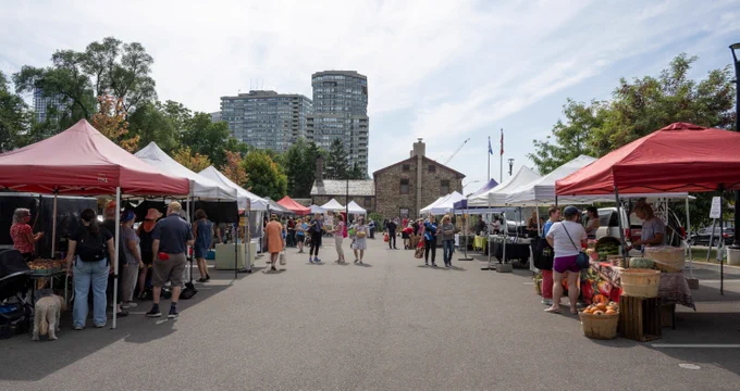 Outdoor market with white and red tents, people browsing stalls, and tall buildings in the background under a partly cloudy sky.