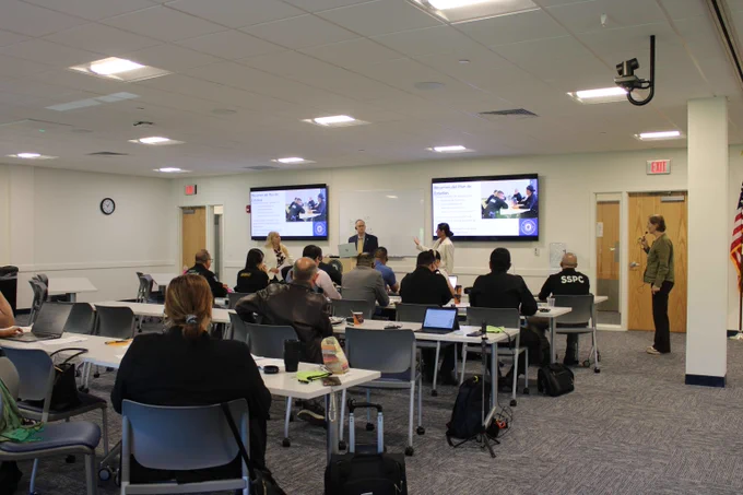 A classroom setting with attendees seated at tables facing presenters and dual screens displaying a presentation slide.