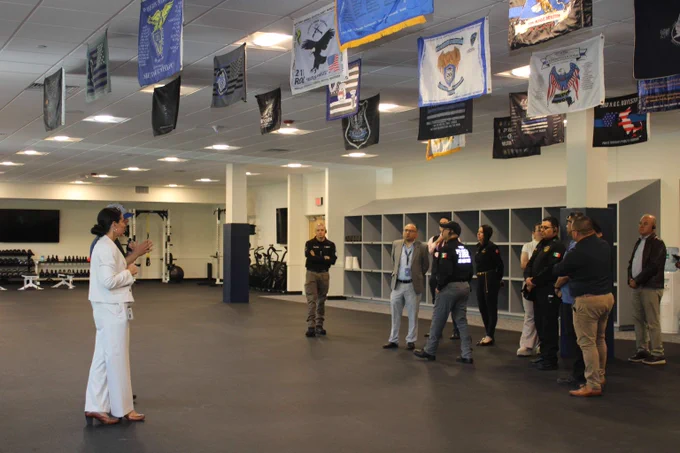 The agencies Deputy Executive Director Captain Amy Fanikos (Retired) speaks to a group Police Officials from Mexico standing in a gymnasium with police academy flags hanging from the ceiling and gym equipment in the background.