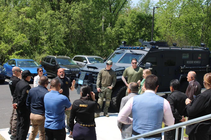 Group of law enforcement officers gathered outdoors near a black armored police SWAT vehicle during a briefing.