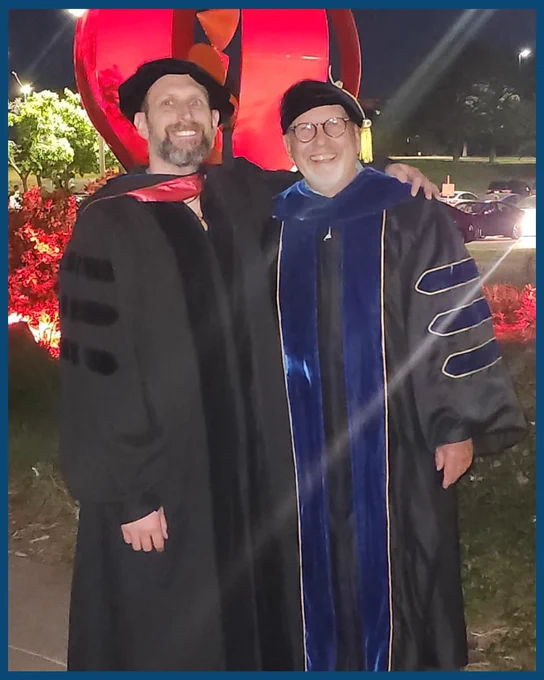 Two people in academic gowns are smiling and posing for a photo together during an outdoor evening event.