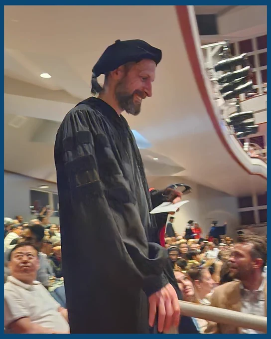 A graduate in ceremonial robes smiles as he walks by a seated crowd in an auditorium, creating a lively and joyful atmosphere.