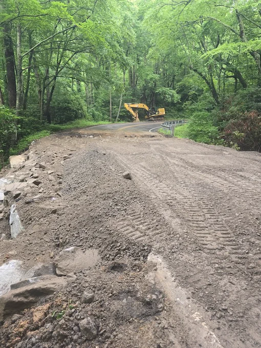 A gravel section of road surrounded by lush green trees, with an excavator parked in the distance. The ground is uneven with visible large rocks and tire tracks.