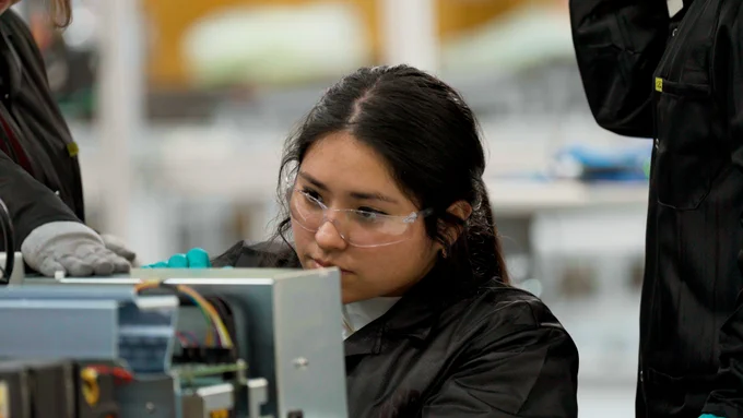 An individual wearing safety goggles and a dark lab coat works intently on a piece of electronic equipment. Others in similar attire are nearby, suggesting a collaborative technical environment.