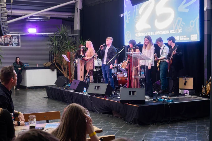 A band performs on an indoor stage with microphones, drums, and guitars. A large screen behind them displays "25" and mentions commercial music. Audience members are seated at tables, watching the performance.