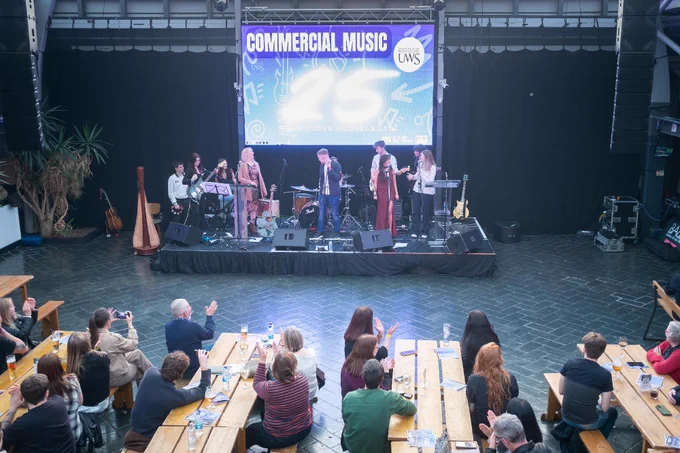 An indoor stage hosts a live music performance with several musicians playing instruments under a large banner reading "Commercial Music 25 UWS." The audience, seated at wooden tables, applauds the performance.