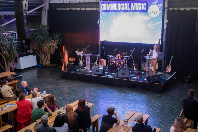 A person is speaking on a stage with a "Commercial Music" banner and a UWS logo. The stage is equipped with musical instruments and microphones. An audience is seated at wooden tables in a spacious hall, listening attentively.