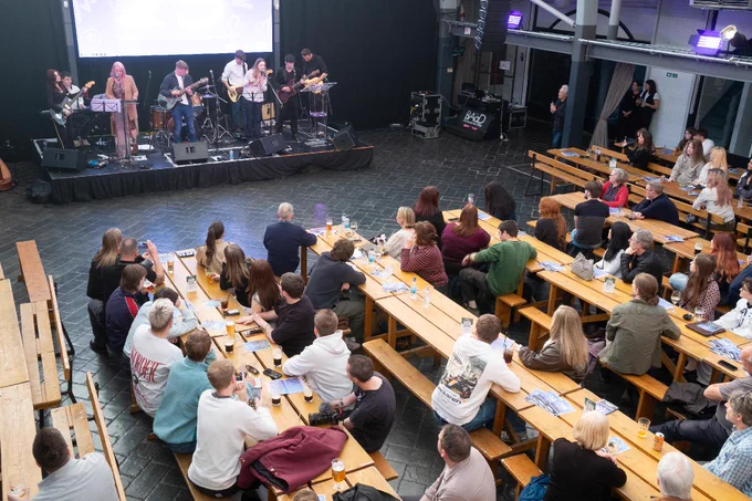 A group of musicians performs on a stage in a large indoor venue. The audience sits at long wooden tables, attentively watching the performance. The atmosphere appears relaxed and engaging. A BBC logo is visible on equipment near the stage.