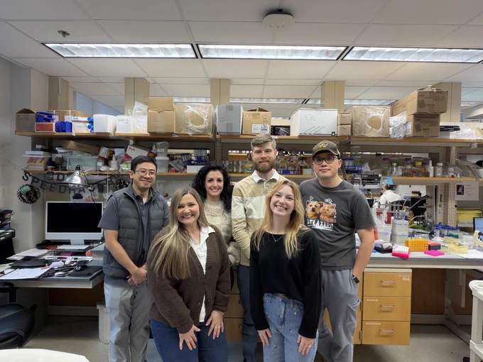 Six members of the Zhu Lab pose together in their research laboratory at Vanderbilt University. The group stands in front of shelving units stocked with laboratory supplies, equipment, and cardboard boxes. From left to right: a man wearing glasses and a gray vest, a woman with long blonde hair in a brown cardigan, a woman with dark curly hair in a beige sweater, a tall man with a beard in a tan and white color-block shirt, a woman with long blonde hair in a black top, and a man wearing glasses and a baseball cap with a graphic t-shirt. Laboratory benches with colorful sample tubes, pipettes, and equipment are visible on the right side of the image.