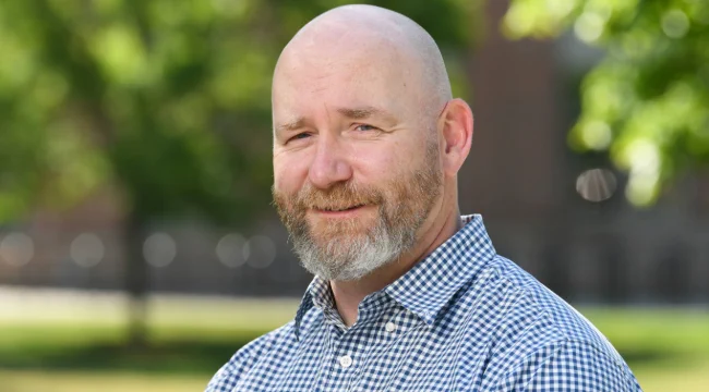 A headshot of a man wearing a blue and white checkered button down outside on UVM's campus.