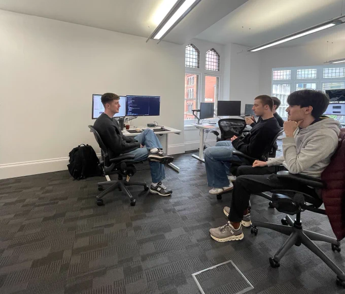 Four software engineers in a bright, modern office gathered for an informal presentation. One team member sits at a white desk with code visible on a dual-monitor setup, turned in his ergonomic chair to speak. Three colleagues sit opposite him in office chairs, listening attentively.